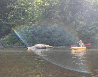 Photo of man and dog on kayak on freshwater lake. A large rock emerges from the water on their right. Rainbow prism over them.