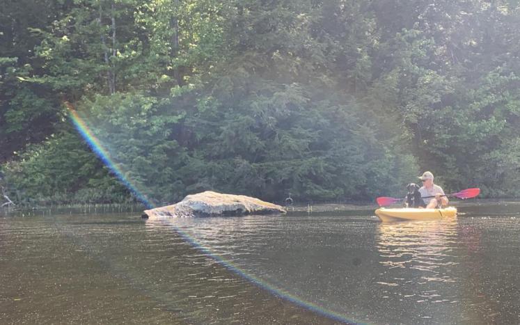 Photo of man and dog on kayak on freshwater lake. A large rock emerges from the water on their right. Rainbow prism over them.